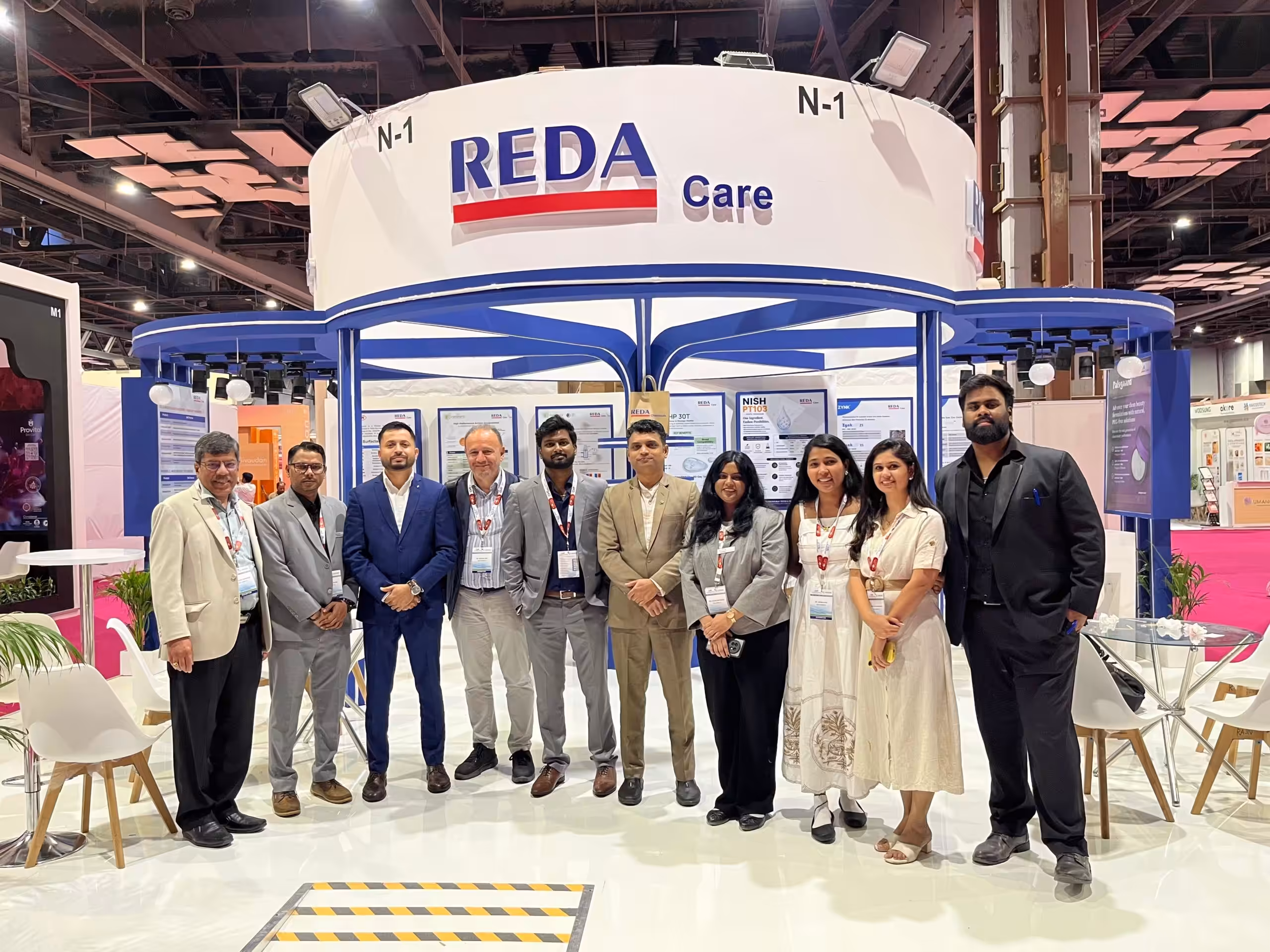 Group of professionals in business attire posing at a REDA Care booth at a trade show venue.